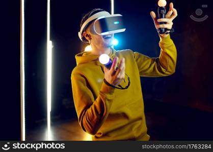 Male gamer plays the game using virtual reality headset and gamepad in luminous cube. Dark playing club interior, spotlight on background, VR technology, 3D vision. Male gamer using virtual reality headset