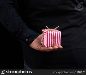 male hand holds a paper-wrapped box with a gift behind his back, man is wearing a black shirt, a concept of surprise