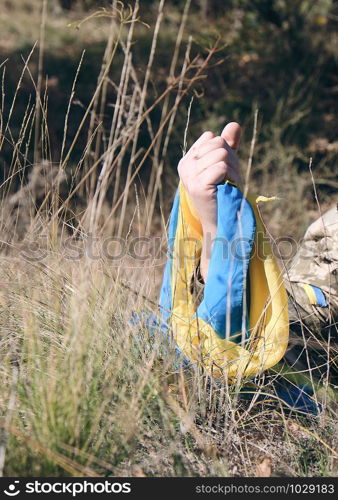 male hand of a Ukrainian army soldier raised up among the grass and holds the state yellow-blue flag, concept of the memory of the dead soldiers