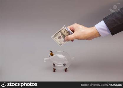 Male hand putting one hundred dollars bill into glass piggy bank isolated on gray