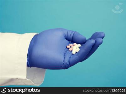 male hand with blue sterile gloves holds white pills, concept of drug treatment for diseases, blue background