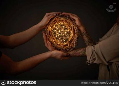 Male hands and woman hands with tattoos holding a bright wreath of twigs with yellow garlands around a black background with copy space for text. Romantic concept. Christmas garland with garlands in male and female hands on a black background with copy space