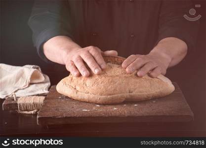 male hands are holding brown baked rye bread over wooden board , black background
