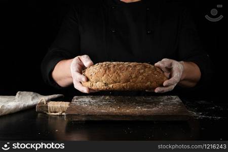 male hands are holding brown baked rye bread over wooden board with flour, black background