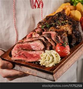 Male hands holding a wooden board with a juicy steak with fragrant butter. Sliced Ribeye Steak with Potatoes, Onions and Baked Cherry Tomatoes.