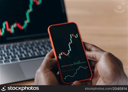 Male hands of African race holds smartphone in front of laptop, broker trader analyzing financial data graphs on phone and computer screen for investment purposes trading charts of stock market. Male hands of African man holds smartphone in front of laptop with trading charts on it