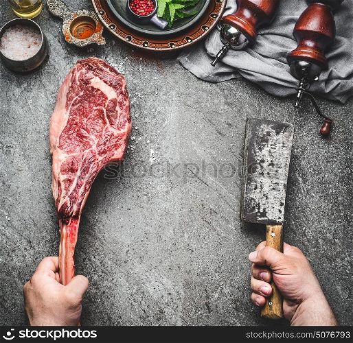 Male hands of butcher or cook holding tomahawk beef steak and meat cleaver on dark rustic kitchen table background with cooking ingredients and condiment, top view, place for text