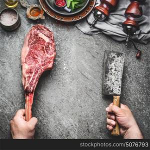 Male hands of butcher or cook holding tomahawk beef steak and meat cleaver on dark rustic kitchen table background with cooking ingredients and condiment, top view, place for text