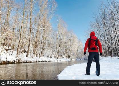 Male hiker enjoying the view of a small river during winter, surrounded by a snowy woods, horizontal frame