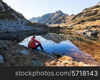 Male hiker takes a rest sitting next a mountain lake. Autumn sunny day. West italian Alps, Oropa, Piemonte, Italy, Europe.