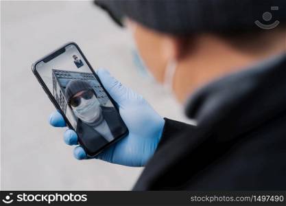 Male holds phone in medical gloves, talks with friend via online conference, greets friend, discuss latest news about coronavirus in their country. Medicine, health care and video consultation
