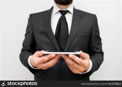 Male human wear formal work suit hold smart hi tech smartphone use hands. Male human with beard wear formal working clothes hold high technology smartphone device. Man dressed in work suit plus tie holding small mobile hi tech phone using two hands