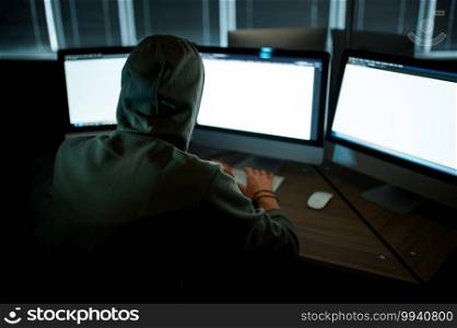 Male internet hacker in hood sitting at monitors, back view. Illegal web programmer at workplace, criminal occupation. Data hacking, cyber security. Male internet hacker in hood sitting at monitors