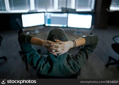 Male internet hacker in hood sitting at screens, back view. Illegal web programmer at workplace, criminal occupation. Data hacking, cyber security. Male hacker in hood sitting at screens, back view