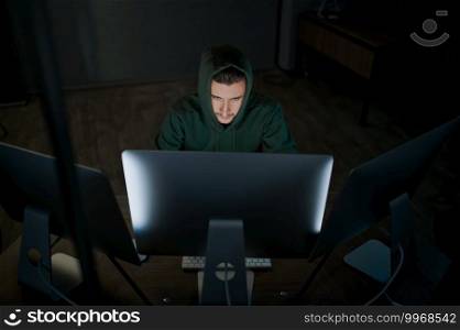 Male internet hacker in hood works on computer, front view. Illegal web programmer at workplace, criminal occupation. Data hacking, cyber security. Male hacker in hood works on computer, front view