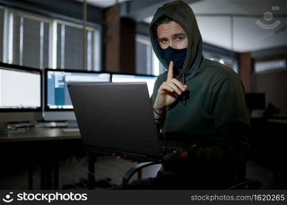 Male internet hacker in hood works on laptop in dark office. Illegal web programmer at workplace, criminal occupation. Data hacking, cyber security. Male hacker in hood works on laptop in dark office