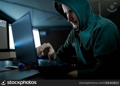 Male internet hacker in hood works on laptop in dark office. Illegal web programmer at workplace, criminal occupation. Data hacking, cyber security. Male internet hacker in hood works on laptop