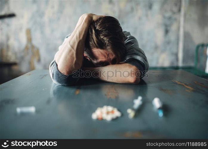 Male junkie sitting at the table with drugs and syringe, grunge room interior on background. Drug addiction concept, addicted people. Male junkie at the table with drugs and syringe