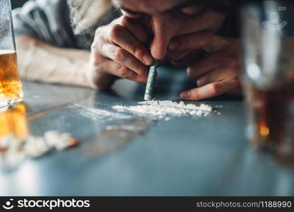 Male junkie sniffing a line of cocaine, grugs and bottle of alcohol on the table on background. Addiction concept, addicted people. Male junkie sniffing a line of cocaine