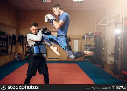 Male kickboxer doing kick in jump, practicing with a personal trainer, workout in gym. Boxer on training, kickboxing practice