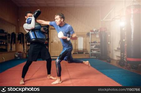 Male kickboxer in gloves practicing hand punch with a personal trainer in pads, workout in gym. Boxer on training, kickboxing practice. Male kickboxer in gloves practicing hand punch