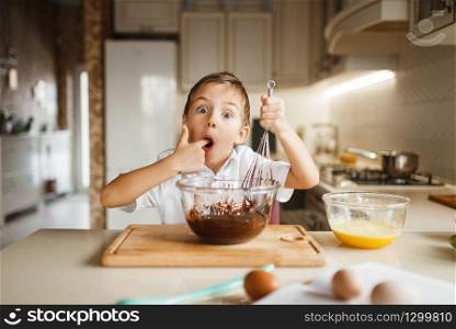 Male kid tastes melted chocolate in a bowl, pastry preparation. Cute boy cooking on the kitchen. Happy child prepares sweet desserts. Male kid tastes melted chocolate in a bowl