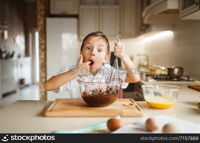 Male kid tastes melted chocolate in a bowl, pastry preparation. Cute boy cooking on the kitchen. Happy child prepares sweet desserts. Male kid tastes melted chocolate in a bowl