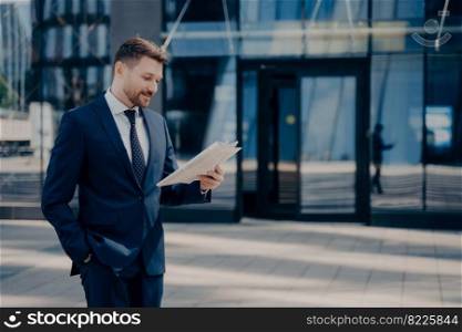 Male lawyer in blue suit reading newspaper, checking out to see if there is anything important while walking alone at office with hand in pocket, young confident businessman enjoying time outdoors. Male lawyer reading newspaper while walking to firm