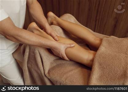 Male masseur pampering legs to young woman in towel, closeup view, professional massage. Massaging and relaxation therapy, body and skin care. Attractive lady in spa salon