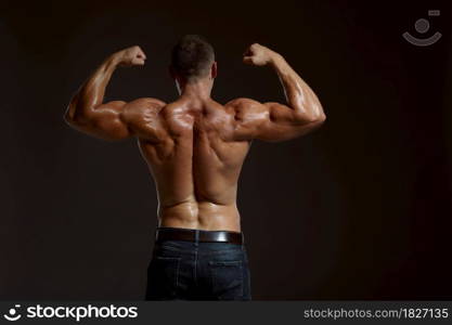 Male muscular athlete poses in studio, back view, dark background. One man with athletic build, shirtless sportsman in jeans pants, active healthy lifestyle. Male muscular athlete poses in studio, back view