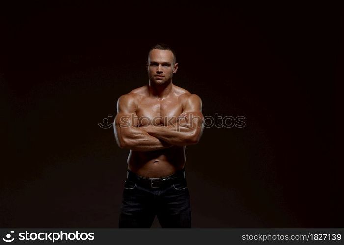 Male muscular athlete shows his power in studio, dark background. One man with athletic build, shirtless sportsman in jeans pants, active healthy lifestyle. Male muscular athlete shows his power in studio