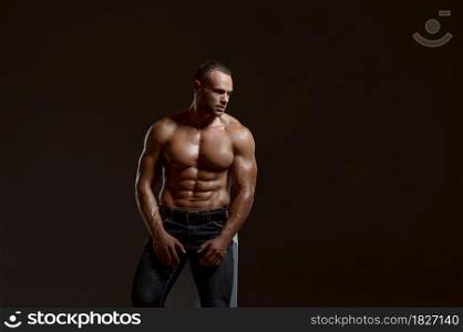 Male muscular athlete sitting on cube in studio, dark background. One man with athletic build, shirtless sportsman in jeans pants, active healthy lifestyle. Male muscular athlete sitting on cube in studio