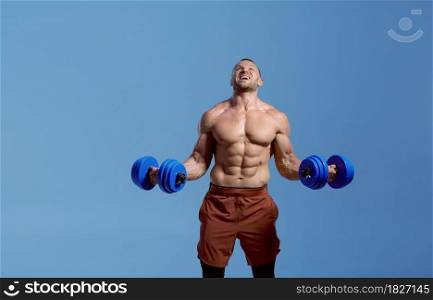 Male muscular athlete with dumbbells poses in studio, blue background. One man with athletic build, shirtless sportsman in sportswear, active healthy lifestyle. Male muscular athlete with dumbbells in studio