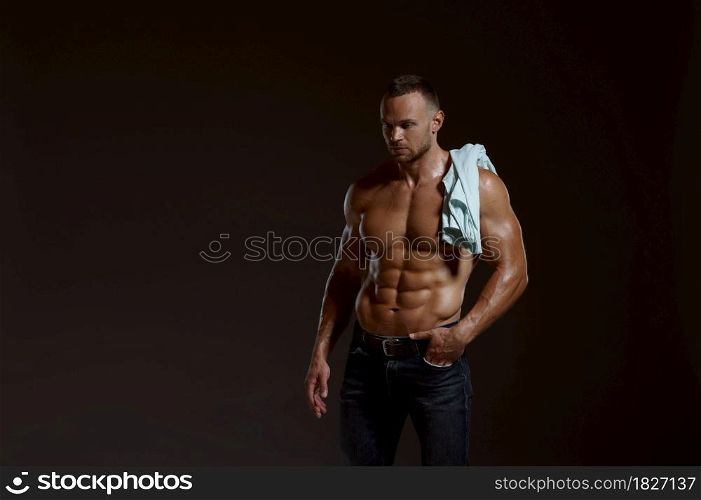 Male muscular athlete with shirt on his shoulder poses in studio, dark background. One man with athletic build, shirtless sportsman in jeans pants, active healthy lifestyle. Male muscular athlete with shirt on his shoulder