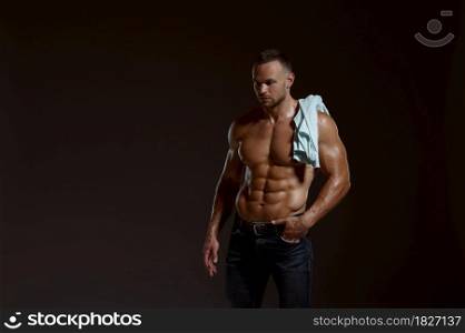 Male muscular athlete with shirt on his shoulder poses in studio, dark background. One man with athletic build, shirtless sportsman in jeans pants, active healthy lifestyle. Male muscular athlete with shirt on his shoulder