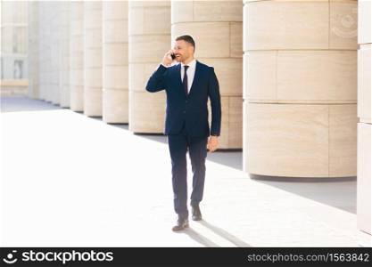 Male office employee calls to someone via modern cellular, wears formal outfit, looks happily aside, has stroll outdoor, focused on something aside, discusses some details of business contract