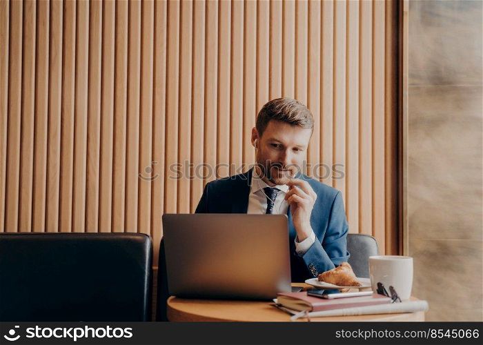 Male office manager wearing blue suit and white shirt, speaking to his workers in online meeting and looking at laptop computer while sitting and working remotely in cafe during business trip. Male office manager during online meeting while sitting in cafe