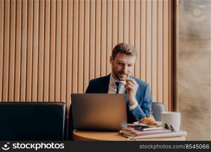 Male office manager wearing blue suit and white shirt, speaking to his workers in online meeting and looking at laptop computer while sitting and working remotely in cafe during business trip. Male office manager during online meeting while sitting in cafe