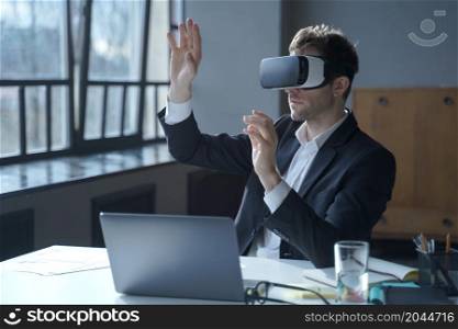 Male office worker in vr headset interacting with digital interface while sitting at desk, touching 3d objects in air, businessman in suit testing virtual reality goggles for business. Male office worker in vr headset interacting with digital interface while sitting at desk