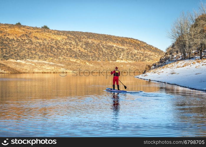 male paddler in red drysuit is paddling a stand up paddleboard on mountain lake in Colorado, winter scenery