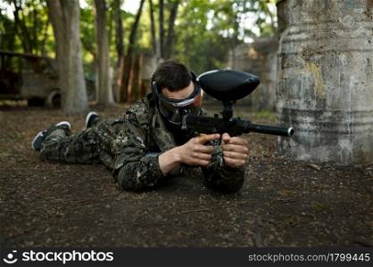 Male paintball player with gun shoots lying down on the ground, playground in the forest on background. Extreme sport with pneumatic weapon and paint bullets or markers, military team game outdoors. Paintball player shoots lying down on the ground