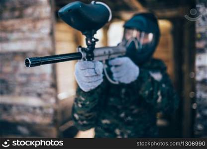Male paintball player with marker gun in hands, front view, focus on weapon, winter battle. Extreme sport game, soldier fights in protection mask and uniform