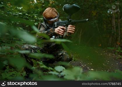 Male paintball warrior with gun shoots from the bushes, playground in the forest on background. Extreme sport with pneumatic weapon and paint bullets or markers, military team game outdoors. Paintball warrior with gun shoots from the bushes
