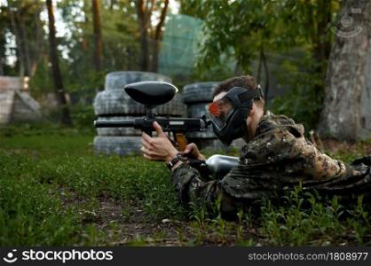 Male paintball warrior with gun shoots lying down on the grass, playground in the forest on background. Extreme sport with pneumatic weapon and paint bullets or markers, military team game outdoors. Paintball warrior shoots lying down on the grass