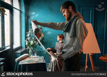 Male painter looking at canvas painting against female poseur in drawing studio. Oil paint, paintbrush drawing