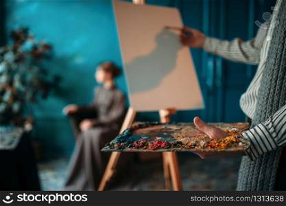 Male painter with palette and brush in hand paints womans portrait on easel. Oil paint, paintbrush drawing. Male painter paints womans portrait