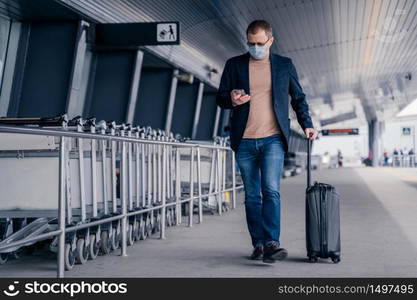 Male passenger walks in airport, holds suitcase, checks newsfeed on smartphone, wears disposable medical mask during pandemic coronavirus, tries to be safe during disease virus. Respiratory symptoms