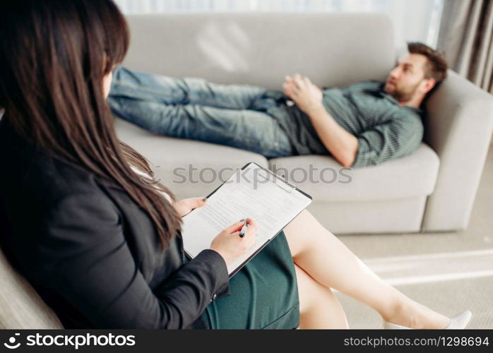 Male patient at psychologist reception, stress. Female doctor writes notes in notepad, professional psychology. Male patient at psychologist reception, stress