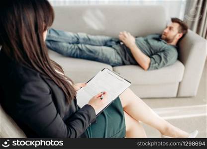 Male patient at psychologist reception, stress. Female doctor writes notes in notepad, professional psychology. Male patient at psychologist reception, stress