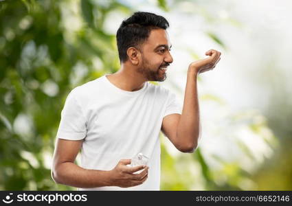 male perfumery, grooming and people concept - happy smiling young indian man smelling perfume over green natural background. happy indian man with perfume